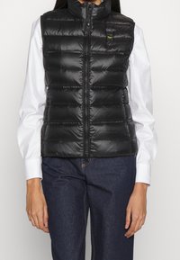 Czarna puchowa kamizelka z wysokim kołnierzem i pionowym przeszyciem. Posiada przedni zamek błyskawiczny oraz małą naszywkę z logo na górnej lewej stronie.