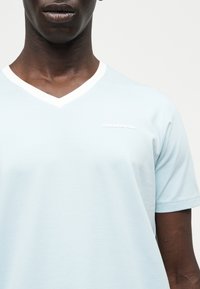 Άτομο με σκουρόχρωμο δέρμα που φοράει ένα γαλάζιο μπλουζάκι V-λαιμόκοψη με λευκή περίγραμμα και μικρό λευκό λογότυπο κειμένου στο αριστερό μέρος του στήθους.