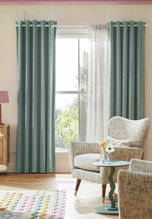 Lichtdurchflutetes Wohnzimmer mit blassblauen Vorhängen, gemustertem Sessel, kleinem rundem Beistelltisch und einer Vase mit Blumen neben einem großen Fenster.