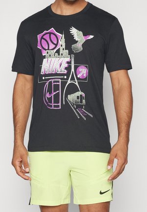 Mężczyzna w czarnej koszulce Nike z różowymi grafikami ptaka, miasta, rakiety tenisowej, metra oraz limonkowych szortach z małym logo Nike.