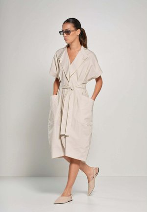 Beige kortjurk met korte mouwen en een wikkelontwerp, vastgebonden in de taille, heeft twee grote zijzakken en een ontspannen silhouet. Gemaakt met lichte ballerina's.