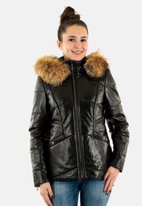 Veste en cuir noire avec capuche bordée de fourrure, dotée d'une fermeture éclair à l'avant et de coutures inclinées. Le mannequin a les mains dans les poches et porte un jean bleu.