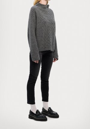 Kvinde iført en løs, grå kabelstrikket sweater, sorte slanke jeans, hvide sokker og sorte chunky loafers, stående mod en ensfarvet baggrund.