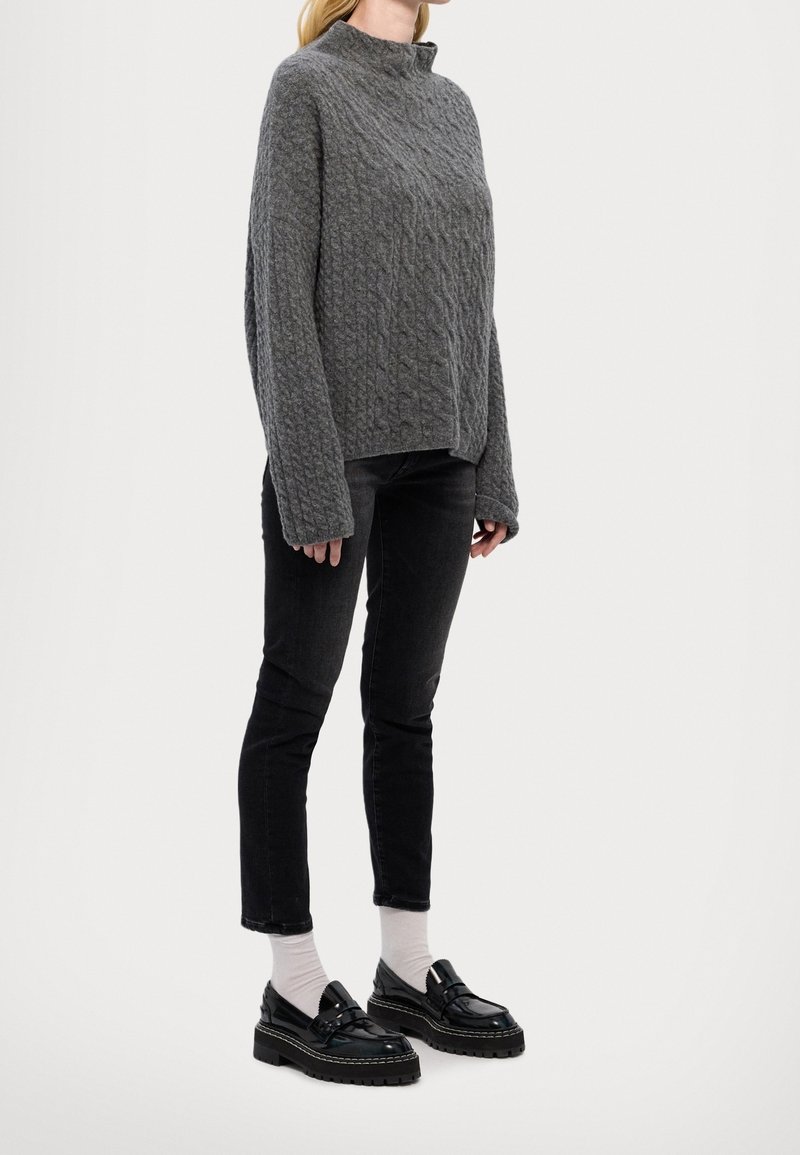 Femme portant un pull gris ample en maille torsadée, un jean slim noir, des chaussettes blanches et des mocassins noirs épais, debout devant un fond uni.