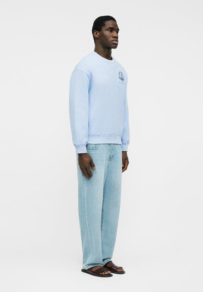 Άνδρας που στέκεται και κοιτά προς τα δεξιά, φορώντας ανοιχτό γαλάζιο φούτερ, ανοιχτό γαλάζιο τζιν και καφέ σανδάλια, σε απλό λευκό φόντο.