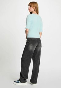 Donna con una maglietta corta e pelosa di colore blu chiaro, jeans neri larghi e scarpe da ginnastica bianche con dettagli blu, in piedi di lato contro uno sfondo neutro.