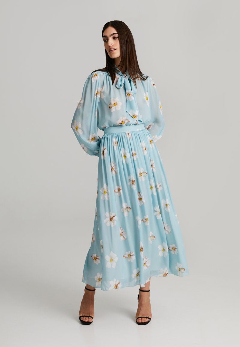 Robe maxi à fleurs bleu clair avec des manches longues et bouffantes, une taille cintrée et un tissu doux et fluide ornés de motifs de fleurs blanches et jaunes.