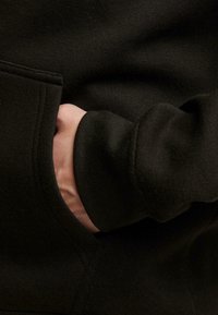 Tejido de sudadera negro con un primer plano de una mano en un bolsillo lateral. El material parece suave y el diseño presenta un puño acanalado.