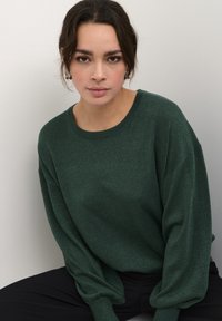 Maglione verde scintillante con una vestibilità morbida, collo rotondo e maniche lunghe. Tessuto liscio, leggero. Pantaloni neri visibili sotto.
