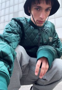 Chaqueta acolchada verde con acentos negros, llevada sobre unos pantalones de chándal grises. La chaqueta cuenta con un alto cuello y costuras detalladas.