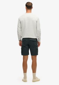 Grå sweatshirt, mørke cargo shorts med sidelommer, beige sokker og lys fodtøj; person set bagfra mod en hvid baggrund.