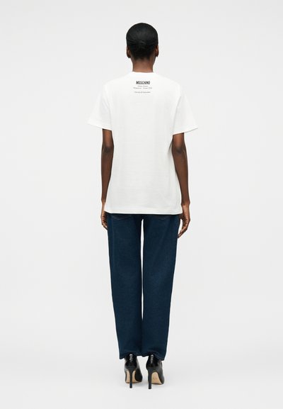 Γυναίκα με σκούρο δέρμα και κοντά μαλλιά, ντυμένη με λευκό t-shirt Moschino, σκούρο μπλε τζιν και μαύρες ψηλοτάκουνες γόβες, φανερή από πίσω.