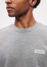 Sweatshirt cinzento de algodão com gola redonda, apresentando um patch de logo texturizado em forma retangular na parte frontal. Acabamento de tecido liso.