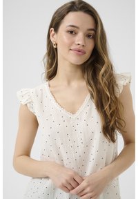 Haut sans manches couleur crème clair avec des pois noirs, coutures à volants aux épaules et encolure en V festonnée. Tissu doux et texturé.