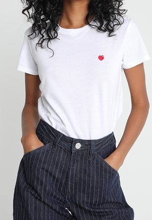 Femme portant une chemise blanche à manches courtes avec un petit cœur rouge sur la poitrine et un pantalon à rayures fines foncé taille haute, les mains dans les poches.