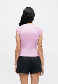 Hosszú fekete hajú nő ujjatlan, világoslila bordázott felsőt és fekete rövidnadrágot visel, háttal áll egy sima fehér háttér előtt.