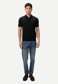 Jeune homme debout, portant un polo noir à manches courtes, un jean bleu et des chaussures marron foncé à lacets sur un fond clair uni.