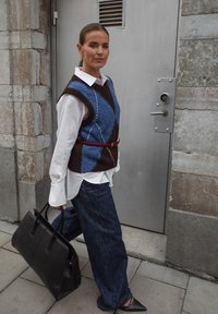 Chaleco de punto en azul y marrón con patrón de diamantes, camisa blanca, jeans oscuros anchos y bolso negro. Zapatos angulares completan el look.