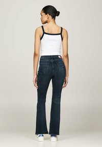 Top sin mangas blanco con ribete azul marino, combinado con jeans acampanados azul oscuro que tienen dos bolsillos traseros y una etiqueta roja, blanca y azul en la cintura.