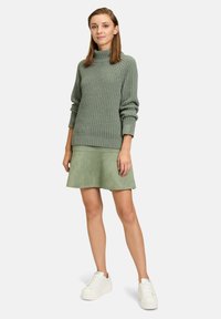 Grüner gestrickter Pullover mit hohem Kragen, kombiniert mit einem passenden grünen Wildlederrock und weißen Sneakern. Strukturierter Stoff, lässige Passform.