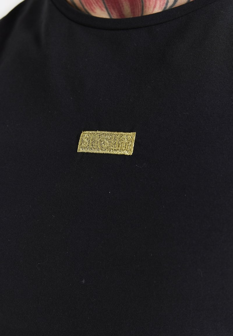 Černé tričko s krátkým rukávem s hladkou texturou a obdélníkovou zlatou vyšívanou nášivkou na hrudi.