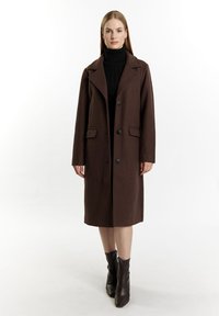 Brun uldfrakke med notched lapel, enkelt knapdesign, to forlommer og fire knapper. Båret over en sort turtleneck.