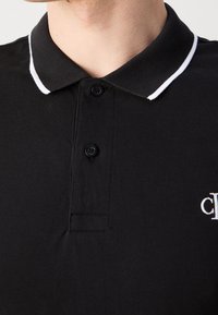 Polo noir avec col bordé de blanc et deux boutons noirs, montrant partiellement un logo brodé blanc sur la poitrine.