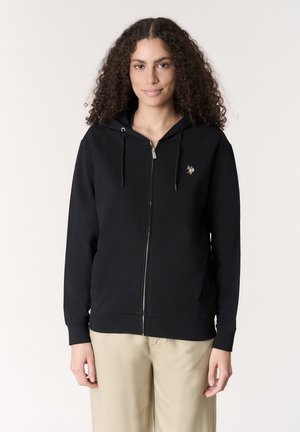 Donna con capelli ricci che indossa una felpa nera con zip e coulisse e pantaloni beige, in piedi davanti a uno sfondo chiaro e semplice.