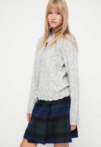 En blond kvinde iført en lyst grå kabelstrikket cardigan og en blå-grønternet nederdel, stående mod en hvid baggrund.