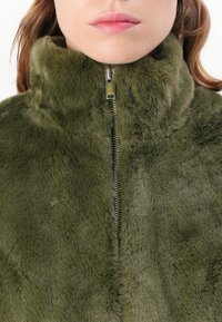 Vrouw in een olijfgroene nep bontjas, ritssluiting omhoog met een hoge kraag, toont het onderste deel van haar gezicht en hals met lichte huid en roze lippen.