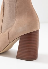 Beżowe zamszowe botki za kostkę z prążkowanym, ciemnym drewnianym obcasem w kształcie bloku oraz elastycznymi bocznymi panelami. Gładka powierzchnia z okrągłym noskiem i przeszyciami.