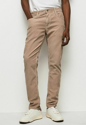 Man draagt beige corduroy broek, witte sneakers en een wit t-shirt, staand met één hand in zijn zak tegen een effen witte achtergrond.