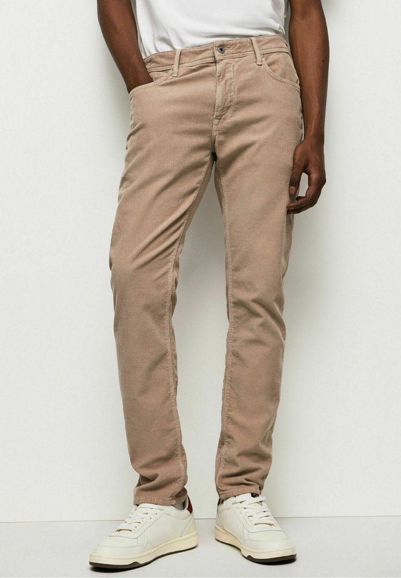 Homme portant un pantalon en velours côtelé beige, des baskets blanches et un t-shirt blanc, debout avec une main dans la poche sur un fond blanc uni.