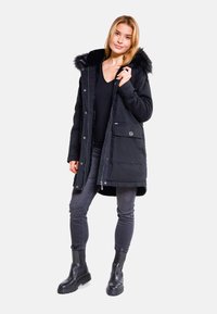 Černá parka s kapucí podšitou kožešinou, zipovými a patentními uzávěry a dvěma předními kapsami. Nosená přes černý top a šedé skinny džíny. Černé kotníkové boty.