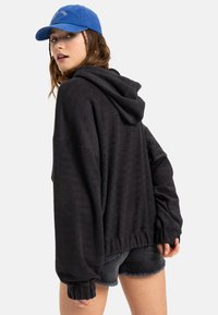 Black, textured hooded sweatshirt with a loose fit and ribbed cuffs, paired with distressed black denim shorts and a blue cap.