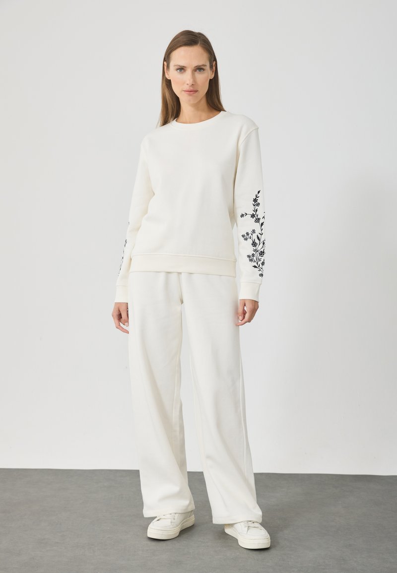 Sudadera de color crema y pantalones de pierna ancha en tejido suave, con bordado floral negro en las mangas. Diseño casual y minimalista.