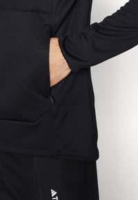 Maglietta sportiva nera con tasca con zip, caratterizzata da una texture liscia e maniche lunghe. La maglietta ha una vestibilità comoda e dettagli di design minimi.