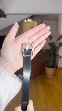 Fekete bőrpánt kézben tartva, ezüst színű négyzet alakú csattal és az élek mentén látható varrással. A háttérben növény látható.