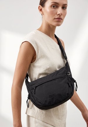 Vrouw draagt een mouwloos beige top en draagt een grote zwarte crossbodytas met rits aan de voorkant en het "GNV"-logo op een lichte achtergrond.