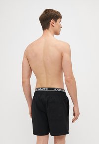Fekete úszónadrág rugalmas derékrésszel, fehér "JACK & JONES" logóval, hátsó zsebbel és sima szövet textúrával.