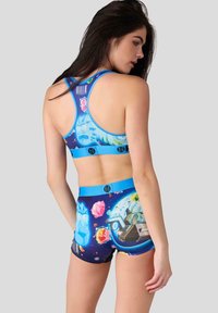 Kvinna i en blå sport-bh och shorts-set med rymdtema, dekorerad med färgglada planeter, kristaller och futuristiska fordon, vänd åt sidan.