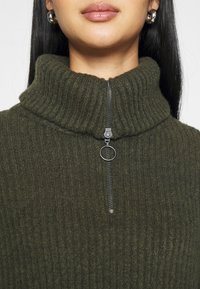 Mørkegrøn, ribbet striktrøje med høj hals og en kvart lynlås; har en sølv lynlås og rundt lynlåshåndtag. Blød tekstur bemærkes.