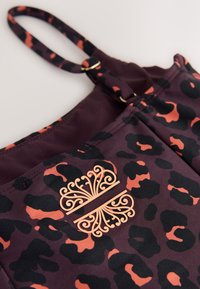 Close-up of dark purple and orange leopard print fabric with adjustable strap and decorative symmetrical orange emblem.