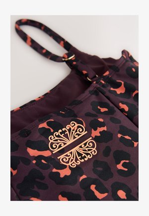 Zbliżenie na ciemnofioletową i pomarańczową tkaninę w cętki lamparta z regulowanym paskiem i ozdobnym symetrycznym pomarańczowym emblematem.