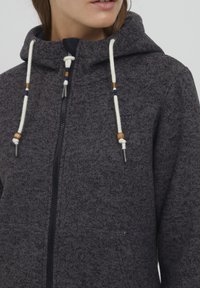Mörkgrå zip-up hoodie i texturerad tyg, med fodrad huva, krämfärgade snören med trätillbehör och en dold framficka.