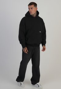 Črna kapuca s sprednjim žepom in izvezenim logotipom; v kombinaciji s temnimi džinsami in belimi športnimi copati. Gladek material, ohlapen kroj.