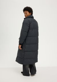 Person with short curly hair wearing a long black puffer coat, dark wide-leg jeans, and black sneakers, standing against a plain background.