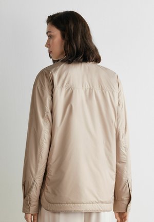 Młoda kobieta z ciemnymi włosami do ramion, ubrana w beżową kurtkę z długimi rękawami, stojąca bokiem na tle gładkiej białej ściany.