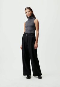 Femme jeune debout portant un pull à col roulé sans manches gris, un pantalon large noir avec ceinture et des chaussures noires sur un fond uni.