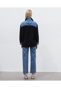 Svart och blå stickad tröja med vadderade axlar, med hög krage, långa ärmar och blå knäppte manschetter. Bärs med blå jeans.